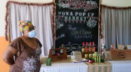 A woman wearing a face mask stands in front of her homemade oils and soaps at a POWA-sponsored market.