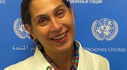 A woman in a yellow suit smiles at the camera in front of a blue and white United Nations sign.