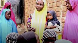 A woman in a yellow hijab holds a baby in her arms as she speaks to other women.