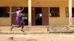 A young girl in a pink shirt and purple dress jumps near a school building.