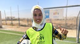 Una joven sonriente sostiene un balón de fútbol en un brazo y da a la cámara un pulgar hacia arriba con la otra mano.