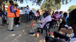 Des membres du personnel de l'ONU se tiennent debout devant un groupe de réfugiés et de demandeurs d'asile assis en groupe, sur des chaises en plastique, à l'ombre d'un arbre, dans le camp de Tongogara. Toutes et tous portent un masque de protection respiratoire.
