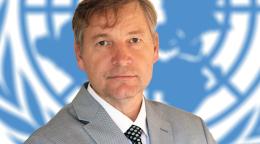 A man, in a grey suit, with his arms folded looks directly at the camera with a United Nations symbol in the background.