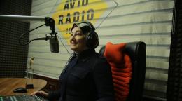 Une femme vêtue d'une tenue bleu foncé et portant un foulard sur la tête et un casque aux oreilles est assise à un bureau, dans un studio radio, devant un microphone et regarde devant elle en souriant.