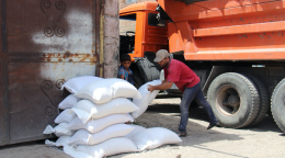 A man in a red shirt unloads white bags from a large orange truck while a small child looks on. 