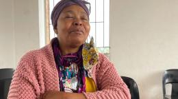 An older woman in a pink sweater sits with her arms folded in a white room. 
