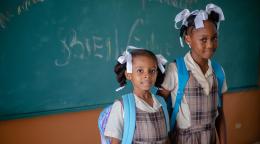 Two girls wearing backpacks and school uniforms stand in front of a chalkboard.