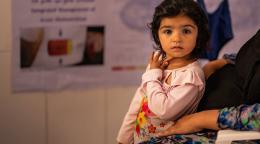 A little girl in a pink shirt looks directly at the camera while she leans on a woman.
