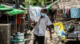 A person in a face mask carries a large white sack on their shoulder down a street with many homes. 