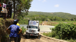 IOM workers stand on the side of the road as a large truck drives down the dirt road and children look down from above.