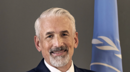 A man with his arms crossed stands in front of the United Nations flag.