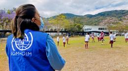 A woman in an IOM vest looks onto a soccer pitch with many kids playing soccer. 