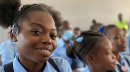 One girl in her school uniform smiles at the camera while the rest of the students stare at the centre of the classroom. 