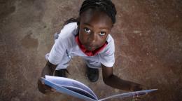 فتاة صغيرة تنظر إلى الكاميرا وتحمل كتابًا مفتوحًا.