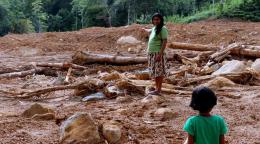 Une femme vêtue d'une jupe mi-longue et d'un t-shirt se tient debout au milieu un champ boueux parsemé de troncs d'arbres déracinés, à la lisière d'une forêt, et parle à un jeune enfant.