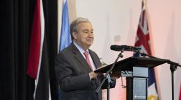 رجل يرتدي بدلة وربطة عنق حمراء، يقف ويتحدث أمام ميكروفون على منصة، وتحيط به مجموعة من الأعلام.