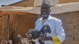 رجل يرتدي قميصًا أزرق ويقف أمام منزل من الطين ويحمل ماعزًا صغيرًا بين ذراعيه وينظر إلى الكاميرا.