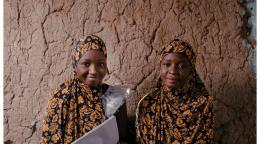 Deux filles qui sDeux jeunes filles de couleur portant un hidjab de couleur noire et jaune sont assies sur un tapis vert, adossées à un mur décrépi, dans une pièce sombre, tiennent chacune un cahier entre les mains et regardent l'objectif en souriant.ourient, sont assises sur le sol contre un mur et se tiennent des dossiers.