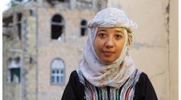 Portrait d'une jeune femme portant un fouloir autour de la tête, les bras croisés, posant devant la caméra, avec en arrière plan un bâtiment délabré.