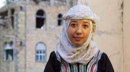 Primer plano de una mujer joven que mira a la cámara con sus brazos sobre su regazo, y en el fondo una edificación en una estado de ruina.