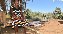 امرأة ترتدي فستانًا تجلس على حافة عربة، في ظل شجرة، بالقرب من حقل، وتنظر إلى الكاميرا ويداها في حضنها.