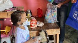 A small girl with a facemask holds a bag of beans while looking at two adults, and in the background there is a table with food on it.
