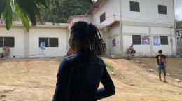 A woman standing with her back to the camera, looking straight ahead, with a building and a yard in the background.