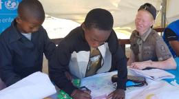 يدرس صبيان صغيران على طاولة تحت خيمة كبيرة أقامتها اليونيسف، بينما طفل ثالث يشاهد بابتسامة كبيرة.