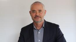 Primer plano de un hombre de pelo corto, barba y bigote que mira a la cámara con seguridad y lleva puesto un traje chaqueta en tono azul oscuro..