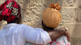 Two women in colorful headwraps embrace, and face away from the camera, in front of a tan-colored cement wall.