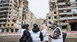 A small group stand before buildings that appear to have been bombed or destroyed.