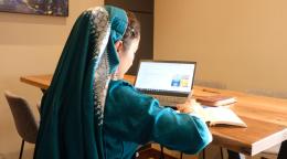 A female in a green satin-like dress with matching headscarf faces away from the camera and works on a laptop.