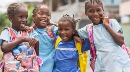 School-age girls in braids and colorful t-shirts and backpacks giggle and embrace, and face the camera.