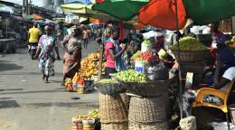 A market scene with men and women walking and surrounded by baskets with vegetables and fruits under umbrellas.