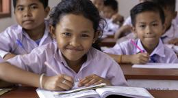 Children in white school uniforms sitting at a desk with books open smiling to camera