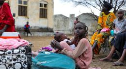 A group of people from Sudan sitting on the ground, the woman at the center is in a pink dress hold
