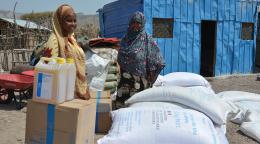 A woman in a brown dress and headscarf stands behind several boxes and cartons and sacks of emergency supplies. A blue cabin is in the background.