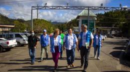 A group of people in different white and blue shirts and vests walk and talk through a grey road where cars are parked