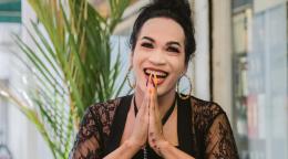 A person in a black shirt folds their hand and smiles while the background has a green plant