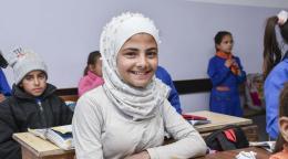 A girl in a white dress and headscarf sits at a school desk with a book open
