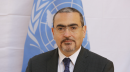 A UN official in a suit and tie poses for a photo in front of a blue UN flag