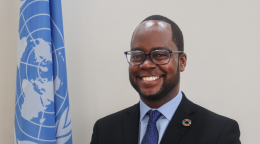 A UN official in a suit and tie smiles and stands next to a UN flag 