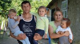 A man and woman stand facing the camera holding their three young children
