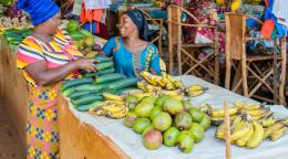 Two women in colourful clothes laugh with each other in a market place, behind a stall selling mangoes and bananas