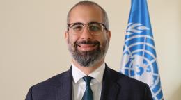 A man in a black suit stands in front of a blue UN flag