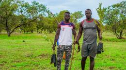 Two men, one in a white shirt and the second in a grey shirt, stand in a green field holding planting tools 