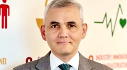 A man in a grey suit and green tie stands in front of a wall painted with the colours of the SDGs