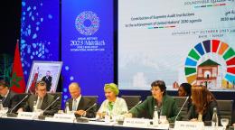 Officials from various countries and institutions on a panel with bright screens and logos behind them.