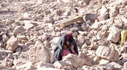 A woman in a red and black dress crouches among the stones, holding her head