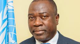 A man in a dark grey suit and blue tie standing next to a blue UN flag.